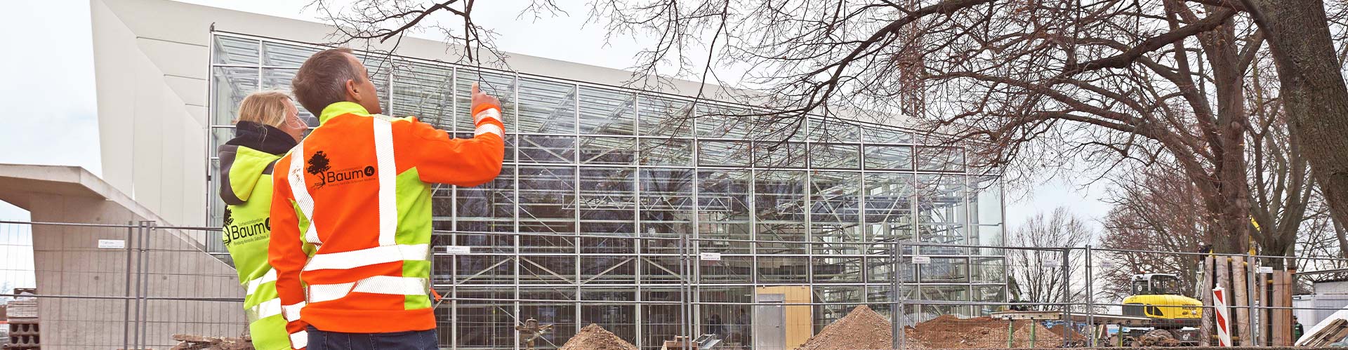 Zwei Mitarbeiter des Sachverständigenbüros Baum 4 GmbH in auffälligen Warnwesten inspizieren einen Baum vor einer modernen Glasfassade und einer Baustelle – symbolisiert professionelle Baumgutachten und Baumpflege im urbanen Raum.